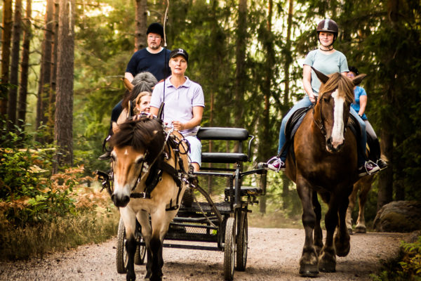 En grupp människor på en skogsväg: en person kör en häst med vagn med passagerare, och en annan person rider en häst bredvid. Höga träd kantar vägen.