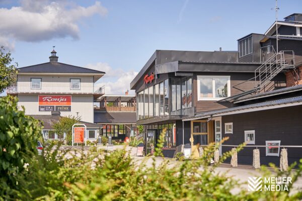 En vy över Ronja Fitness & Relax i Vimmerby, som visar flera svarta och ljusa byggnader med stora fönster och en logotyp synlig på en av fasaderna. I förgrunden syns buskage och i bakgrunden en del av Ronja Hotel.