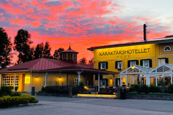 Ett gult hotell med texten "Karaktärshotellet" på fasaden, ett rött tak och en inglasad entré, fotograferat i solnedgången med en dramatisk röd himmel.