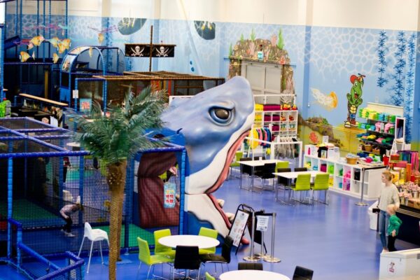 En stor och färgglad lekplats inomhus med en jättelik hajfigur, klätterställningar med piratskeppstema och flera bord och stolar. I bakgrunden finns en butiksdel med hyllor fulla av varor, och väggarna är målade med undervattensmotiv.