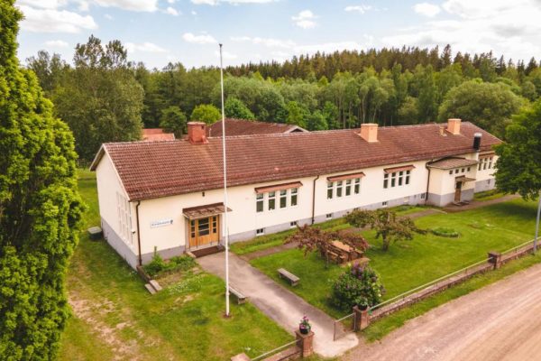 En luftbild av en större, ljusgul byggnad med rött tak, omgiven av en grön gräsmatta och träd. En flaggstång står framför byggnaden, och en grusväg leder fram till den.