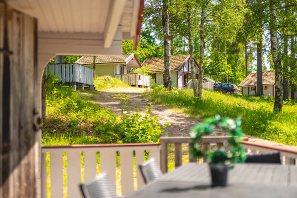 En veranda med ett utomhusbord och stolar, som vetter mot en grusväg med flera stugor och träd i bakgrunden.