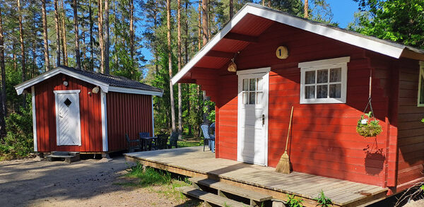 Två små röda stugor med vita knutar och dörrar står framför en tallskog, med en av stugorna med en liten träveranda.