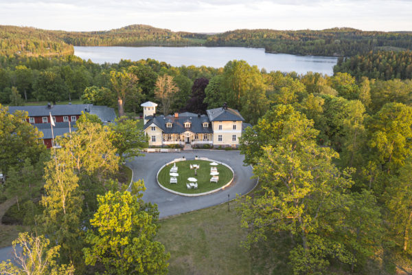 Flygbild över herrgård med gul fasad och mörkt tak, omgiven av skog och sjö. Framför finns en uppfart med vita möbler.