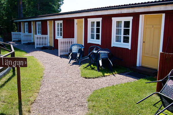 En rad med små, röda stugor med vita fönsterkarmar och gula dörrar, framför vilka det finns grusgångar, gräsmattor och några utemöbler. En träskylt med texten "Längan" syns i förgrunden.