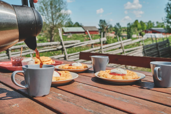 En hand häller kaffe från en termos i en grå mugg på ett rustikt träbord. På bordet står även tallrikar med våfflor toppade med grädde och sylt, samt fler kaffekoppar. I bakgrunden syns ett pittoreskt landskap med röda stugor och ett trästaket.