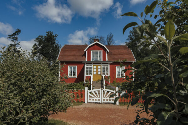 Ett rött trähus med vita knutar och gul dörr, omgivet av grönska under en blå himmel med moln.
