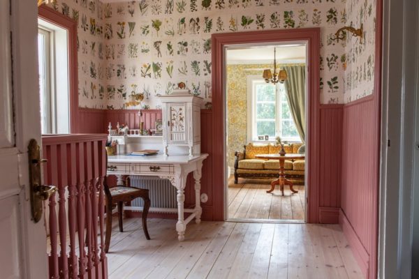 En rumsvy med ljusa trägolv och tapeter med botaniska motiv ovanför rosa träpaneler. I mitten finns ett vitt skrivbord med en vit byrå och en stol. Till höger leder en öppen dörröppning in till ett annat rum med en soffa och ett fönster. Till vänster syns en del av en vit dörr med mässingshandtag och ett rosa räcke.