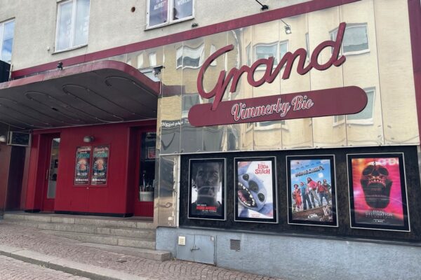 En bild på Vimmerby bios entré med en röd skylt med texten Grand Vimmerby bio. Det är en metallvägg med röda dörrar och fyra filmplanscher.
