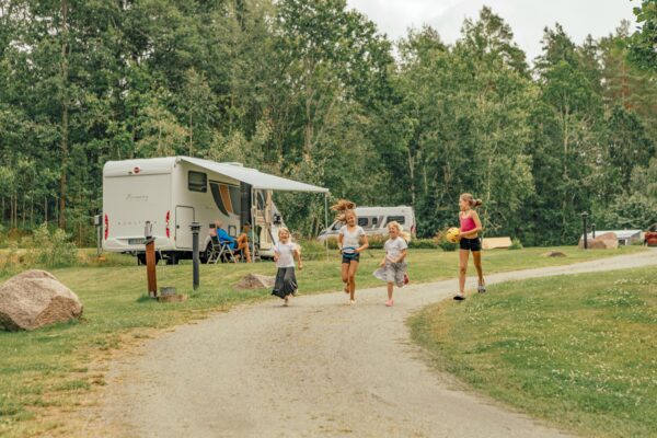 Fyra flickor springer glatt längs en grusväg på en campingplats. I bakgrunden står en husbil och det finns träd och gräs.