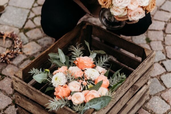 En trälåda fylld med buketter av rosa, persikofärgade och vita rosor blandat med gröna blad. I bakgrunden håller en person en liknande bukett. Allt står på ett gatstensgolv.