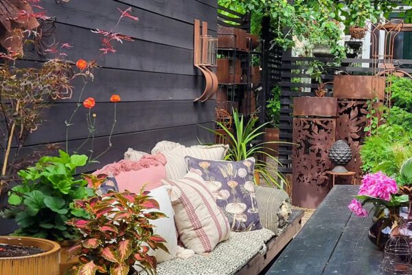 En mysig utomhusplats med en soffa fylld med kuddar, omgiven av växter och dekorativa metallföremål, intill en mörk trävägg.