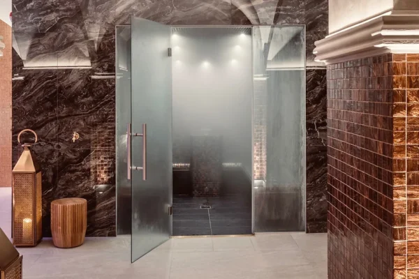 En modern ångbastu med glasdörr, mörka marmorväggar och ett gyllene kaklat pelare.