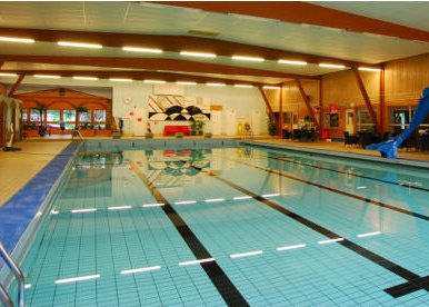 En rymlig inomhuspool med flera banor och en vattenrutschkana i Vimmerby. Taket har synliga träbalkar och är utrustat med radvis av lampor. I bakgrunden syns ett annat poolområde och en entré.