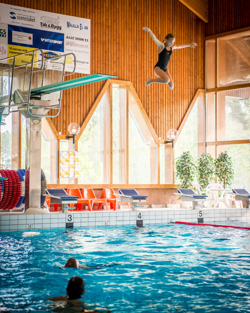 En ung tjej i svart baddräkt hoppar elegant från en trampolin ner i en simbassäng i Vimmerby simhall. I vattnet syns delar av andra simmare. Bakom trampolinen finns en vägg med reklam för olika företag.