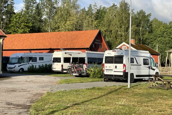 Fyra husbilar står parkerade vid en röd ladugård i lantlig miljö med omgivande grönska och träd.