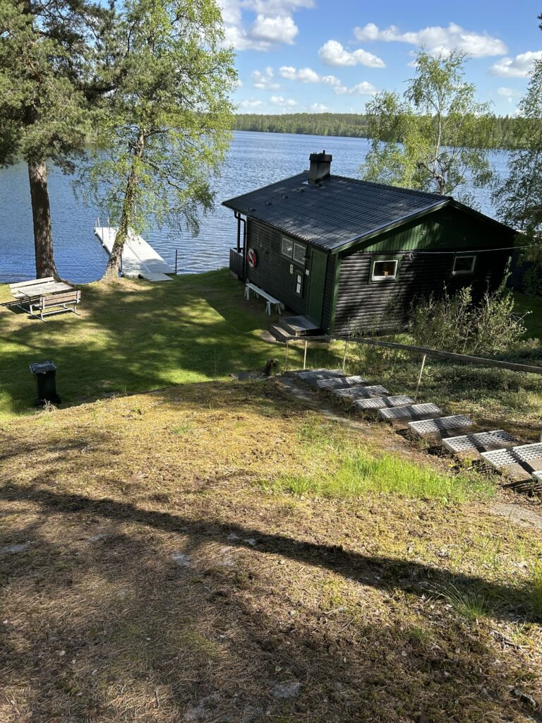 En mörk stuga vid en sjö i Vimmerby, med en brygga som sträcker sig ut i vattnet. En stig med trästeg leder ner till stugan från en gräsbevuxen sluttning. Träd omger stugan och sjön, och himlen är molnig.