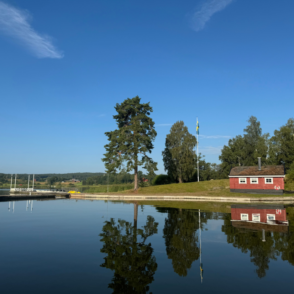 En fridfull scen vid en sjö i Vimmerby, med en liten röd stuga och en brygga på den motsatta stranden. En svensk flagga vajar från en flaggstång, och träd speglar sig i det lugna vattnet under en klarblå himmel.