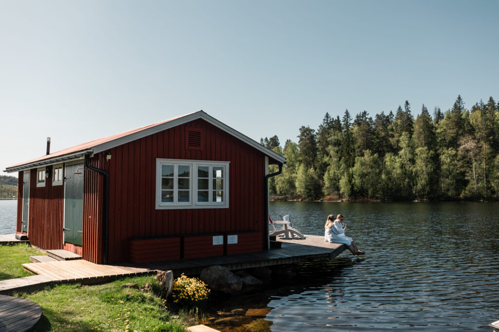 En röd stuga med vita fönster, med en träbrygga som sträcker sig ut över en lugn sjö. En person sitter på bryggan och doppar fötterna i vattnet, med en grön skog i bakgrunden.