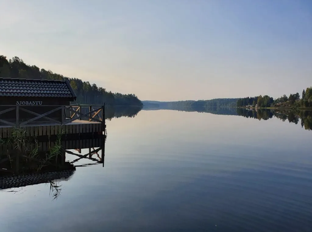 En fridfull sjö i Vimmerby, med en Sjöbastu med träterrass vid strandkanten. Skogsklädda stränder ramar in sjön, vars yta reflekterar den ljusa himlen.