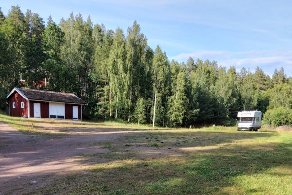 Husbil parkerad vid skogsbryn bredvid en liten röd byggnad, omgiven av gräs och grusplan i lantlig miljö.