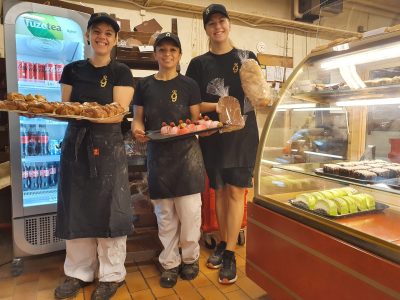 Tre glada bagare, klädda i svarta t-shirts med en logotyp, svarta förkläden och kepsar, står i ett bageri och håller upp brickor med nybakat bröd och bakverk. I bakgrunden syns en kyl med drycker och en displaymonter med fler bakverk.