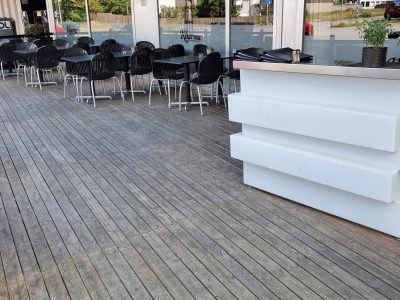 En stor uteservering med trädäck, svarta bord och stolar framför en restaurang med stora fönster. En vit bar eller receptionsdisk står till höger, och i bakgrunden syns träd och byggnader.