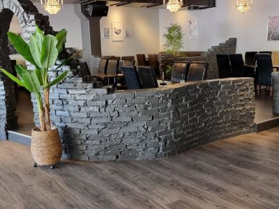 En modern restaurang med mörkt trägolv, mörka väggar, ett stort palmträd, svarta bord och stolar, samt väggar och valv i mörkgrå stenimitation med kristallkronor.