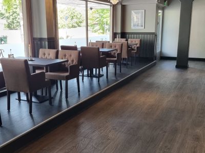 En elegant restauranginteriör med mörkt trägolv, stora fönster och en upphöjd del med bord och stolar. Rummet är inrett med bruna stolar, mörka bord och hängande kristallkronor.