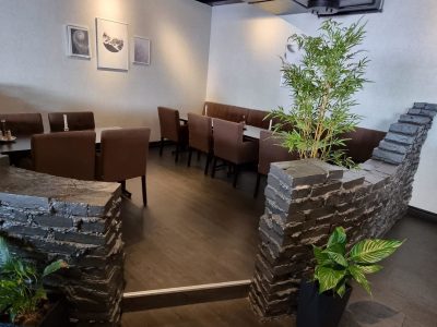 En modern restauranginteriör med mörkt trägolv, vita väggar dekorerade med tavlor, och mörka bord och stolar. Rummet har också inredning av mörk stenimitation och gröna växter som bambu och andra krukväxter.