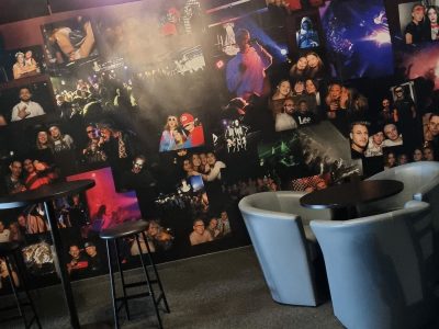 En mörk bar eller lounge med en fotovägg, höga barbord, vita fåtöljer och en glittrig ljuskrona.