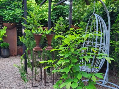En frodig trädgård med en grusgång, omgiven av olika växter och träd. I bakgrunden finns en pergola, dekorativa urnor på stativ och en hängande äggformad stol.