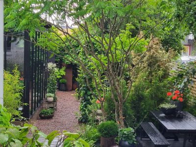 En mysig trädgård med en grusgång, lummiga växter, och en mörk utemöbelgrupp på ett trädäck.