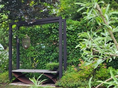 En trädgård med en sittbänk under en svart pergola. Bänken har en distinkt bas av metallvalv. Bakom bänken växer en tät murgröna, och runtomkring finns lummiga träd och buskar. Ett dekorativt hängande lykta pryder pergolan.