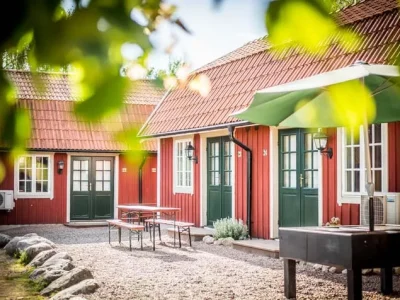 En utomhusbild av två röda stugor med tegelröda tak och gröna dörrar. Mellan dem finns en grusplan med en uteplats under en grön parasoll.