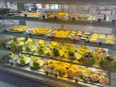 Ett välfyllt kyldisk med en mängd olika bakverk och kakor, inklusive prinsesstårtor, cheesecakes och småbröd, presenterade på hyllor. I bakgrunden syns burkar med "PÄRONKONJAK" och andra förpackningar.