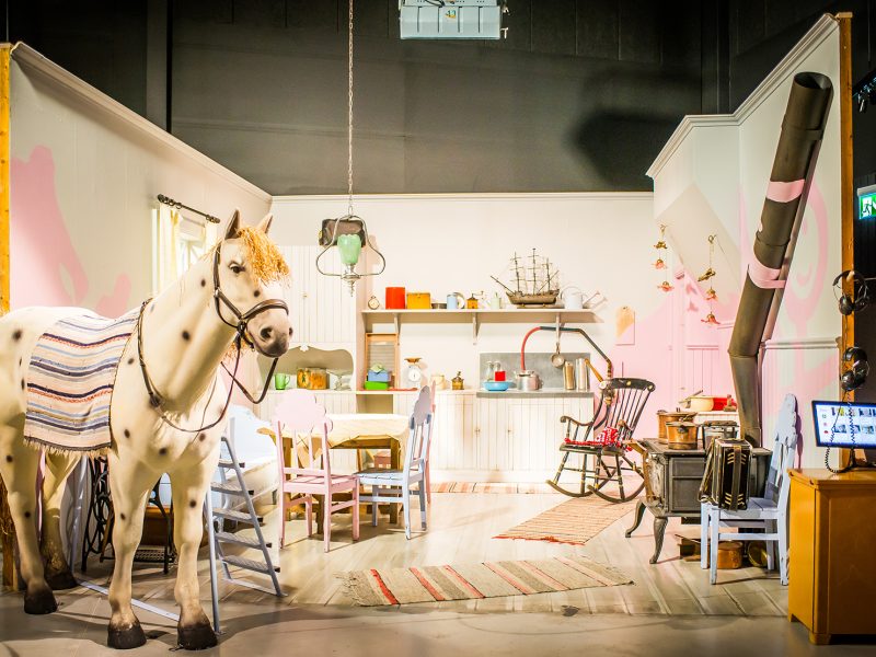 En inomhusscen som ser ut som en filminspelningsplats eller museumutställning. I förgrunden står en vit häst med svarta prickar och en filt. Bakom hästen finns ett rum möblerat som ett kök med ett bord, stolar, hyllor med köksredskap och en vedspis