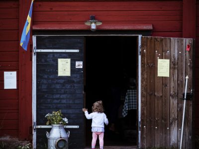 En liten flicka i rosa byxor och vit tröja står vid ingången till ett rött hus med en skylt som säger "Café Sörgården". Dörren är öppen och en svensk flagga syns till vänster. En gammal mjölkkanna med blommor står vid ingången.