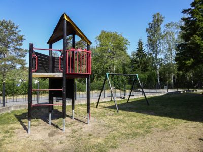 En lekplats med en klätterställning i trä och metall med ett gult tak och röda räcken, bredvid en gunga med två säten. Lekplatsen ligger på en gräsmatta som delvis är torr, omgiven av träd och en svart metallstaket under en klarblå himmel.