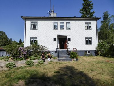 Vit tvåvåningsvilla med trappa upp till entrén, omgiven av grönska och blommor. Två personer sitter i skuggan vid en rabatt framför huset.