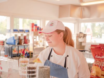 En person i vit kockrock, blått förkläde och vit keps tittar neråt med ett leende, stående bakom en disk i en butik eller servering.
