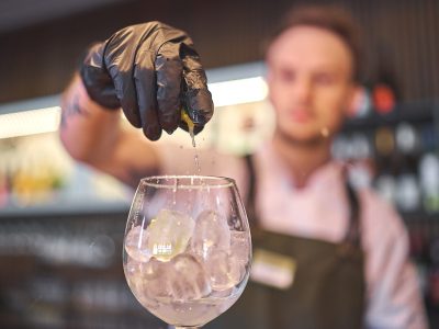 En bartender i svarta handskar pressar en limeklyfta ner i ett glas med isbitar och dryck.