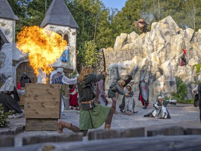 En spännande scen med en stor explosion av eld i förgrunden, som en del av en utomhusföreställning av bröderna lejonhjärta. Flera aktörer i historiska dräkter reagerar på elden, med en medeltida bykuliss och klippformationer i bakgrunden.