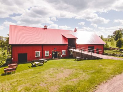 En stor röd lada med ett matchande rött plåttak står på en grön gräsmatta under en delvis molnig himmel i Vimmerby. Utanför finns flera picknickbord, och en ramp leder upp till en av dörrarna på ladans baksida.
