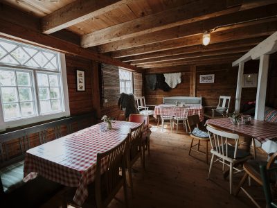 En mysig, rustik restaurang eller ett kafé i Vimmerby med träpaneler på väggarna och i taket. Borden är dukade med röd- och vitrutiga dukar, och olika stolar är utplacerade. Ett stort fönster släpper in naturligt ljus.
