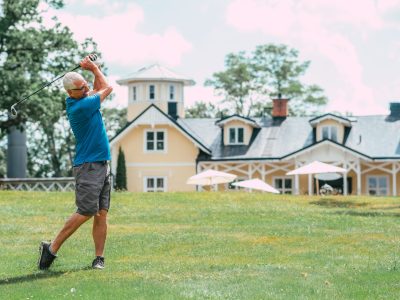 En man i blå pikétröja och shorts svingar en golfklubba på en grön golfbana med en stor gul byggnad i bakgrunden.