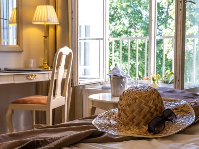Ett ljust hotellrum med en stråhatt och solglasögon på sängen i förgrunden, ett skrivbord till vänster, och ett fönster som visar grönska utanför. En champagnekylare och glas står på ett litet bord vid fönstret.