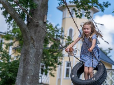 En flicka med ljusa flätor och en vit rosett i håret ler medan hon står på en gunga gjord av ett däck, upphängd i en stor trädgren. I bakgrunden syns en gul byggnad med ett torn.