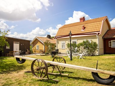 En stor, gammaldags gungbräda med trähjul på en gräsmatta, framför traditionella svenska hus med röda tak.