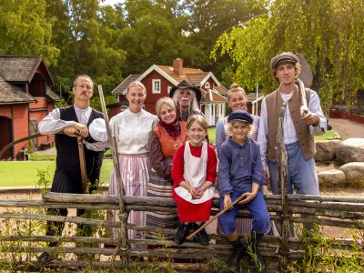 Emil i Lönneberga står vid gärdsgården tillsammans med sin familj och andra figurer. Han har blåa kläder, blå mössa och håller i en trä bysse.